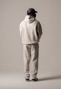 Mężczyzna odwrócony tyłem, ubrany w beżowy hoodie z napisem "BLKVIS", dopasowane spodnie dresowe, czapkę w kolorze czarnym oraz czarno-białe sneakersy na jednolitym tle.