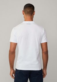Camiseta de algodón blanca con mangas cortas, con un cuello redondo y un sutil logo negro "HACKETT" en la parte posterior. Textura suave.