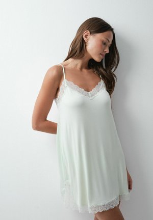 Femme portant une robe nuisette blanche sans manches avec un bord en dentelle au niveau de l'encolure et de l'ourlet, debout contre un mur blanc uni.