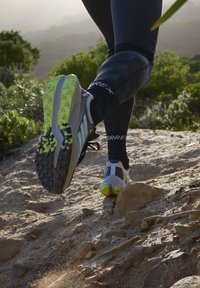 Zapatillas de correr con parte superior gris, acentos negros y suela amarilla neón. Patrón de superficie diseñado para agarre en terrenos irregulares.