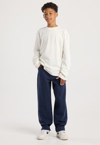 Poika seisoo kädet taskuissa päällään valkoinen pitkähiha paita, tummansiniset housut ja valkoiset tennarit, joissa on musta swoosh-logo.