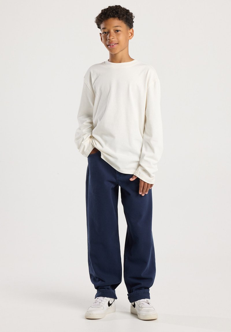 Poika seisoo kädet taskuissa päällään valkoinen pitkähiha paita, tummansiniset housut ja valkoiset tennarit, joissa on musta swoosh-logo.