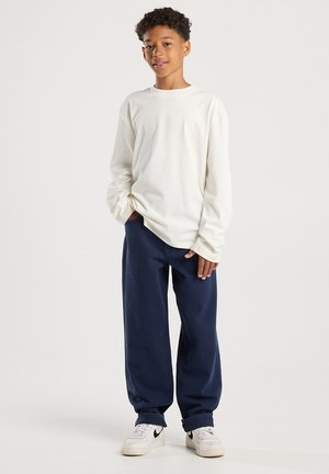 Ragazzo in piedi che indossa una camicia bianca a maniche lunghe, pantaloni larghi blu scuro e scarpe da ginnastica bianche con logo Nike nero su sfondo bianco.
