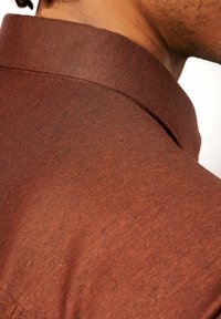 Details van de kraag van een bruine linnen shirt, met een gestructureerde stof en een subtiel ruitpatroon, en een nette afwerking van de randen.