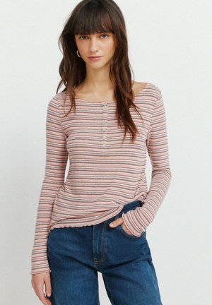 Nuori nainen, jolla on päällään vaaleanpunaraitainen pitkähihainen paita nappeineen ja siniset farkut, seisoo käsi yhdessä taskussa valkoista taustaa vasten.