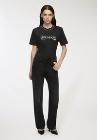 Czarny t-shirt z napisem "MAGNIFIQUE" w kolorze białym, połączony z czarnymi dżinsami o wysokim stanie. Modelka stoi w eleganckich czarnych szpilkach.