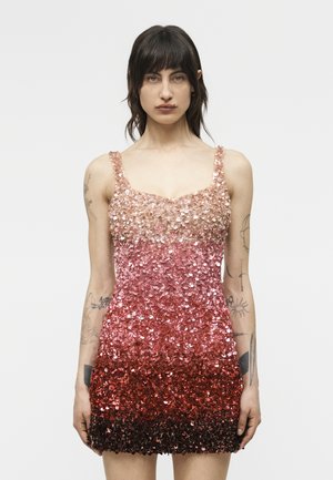 Žena s tmavými vlasmi, oblečená v bezrukávových ombré flitrovaných šatách prechádzajúcich z svetloružovej do tmavočervenej, stojaca na obyčajnom pozadí.