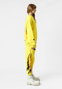Sudadera amarilla y pantalones de chándal a juego con un detalle de flecos y un patrón de alas negras. Botines blancos completan el atuendo.