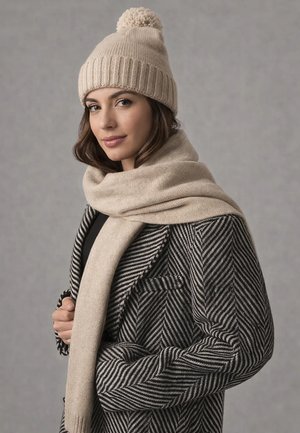 Woman wearing a beige knit hat with pom-pom, matching scarf, and black-and-white herringbone patterned coat, posing against a gray background.