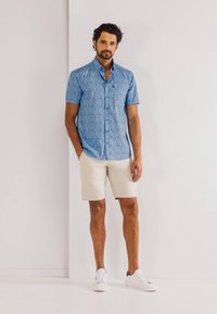 Lichtblauwe short-sleeve shirt met bloemenpatroon, gecombineerd met beige shorts en witte sneakers, tegen een effen witte achtergrond.