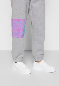 Pantalones deportivos gris claro con un parche gráfico vibrante en rosa y azul en la parte baja de la pierna, complementados con zapatillas blancas con detalles texturizados.
