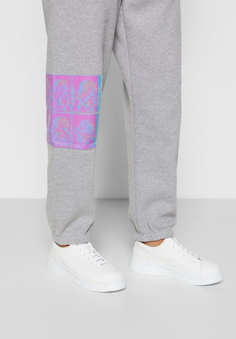 Pantalones deportivos gris claro con un parche gráfico vibrante en rosa y azul en la parte baja de la pierna, complementados con zapatillas blancas con detalles texturizados.