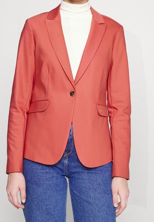 Persona que lleva un blazer coral con un solo botón sobre un cuello alto blanco y vaqueros azules, de pie frente a un fondo liso.