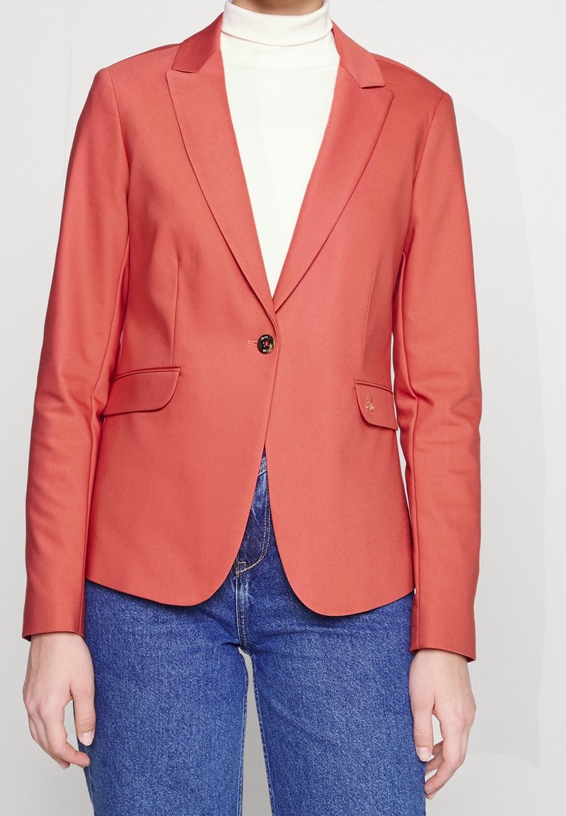 Personne portant un blazer corail à un seul bouton sur un col roulé blanc et un jean en denim bleu, debout devant un fond uni.