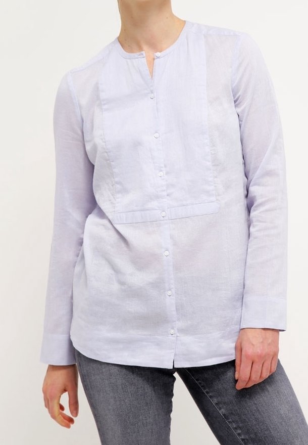 Femme portant une chemise bleu clair à manches longues boutonnée avec un col mandarin et un jean gris foncé, debout devant un fond uni.