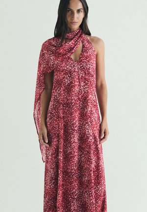 Mujer con vestido rojo sin mangas con patrón de manchas rosas y tela a juego que cae por un hombro sobre un fondo liso.
