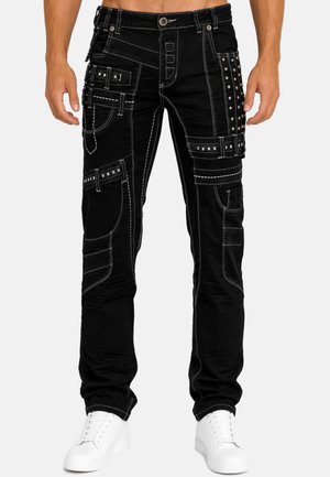 Man wearing black cargo pants with white stitching, metal studs, and multiple pockets paired with white sneakers on a light background.