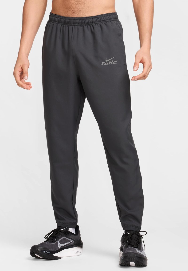 Harmaat urheiluhousut, joissa on joustava vyötärönauha ja Nike-logo vasemmassa reisissä. Materiaali näyttää sileältä, ja housuissa on kapeneva malli. Mustat lenkkitossut näkyvissä.