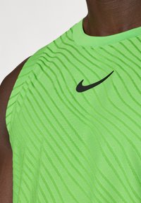 Πράσινη αθλητική τοπ χωρίς μανίκια με υφή κυματοειδούς σχεδίου και μια μαύρη λο γότυπο της Nike στο στήθος. Λείος ύφασμα με στρογγυλή λαιμόκοψη.