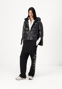 Black puffer jacket with a fitted design, shiny texture, and a hood, paired with loose black pants featuring white graphic detail, and light sneakers.