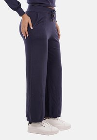 Persona che indossa pantaloni sportivi larghi blu navy con vita regolabile e sneakers bianche, in piedi contro uno sfondo chiaro e semplice.