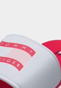 Κοντινό πλάνο λευκής σαγιονάρας με κόκκινη σόλα που φέρει το κόκκινο και ροζ λογότυπο Tommy Hilfiger στο λουρί.