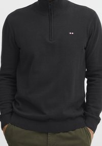 Pull noir à fermeture éclair avec un col montant. Comprend des manches longues, des poignets et un ourlet côtelés, ainsi qu'un petit logo tricolore sur la poitrine. Texture douce.