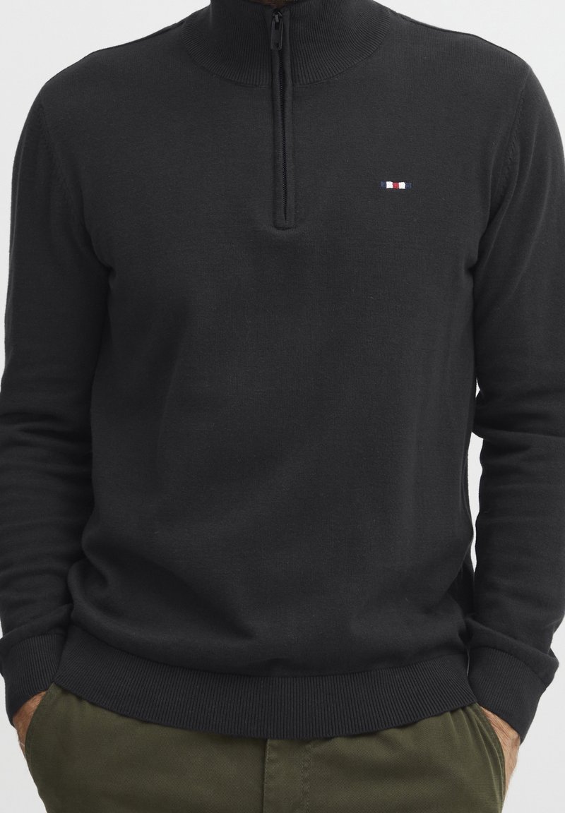 Pull noir à fermeture éclair avec un col montant. Comprend des manches longues, des poignets et un ourlet côtelés, ainsi qu'un petit logo tricolore sur la poitrine. Texture douce.