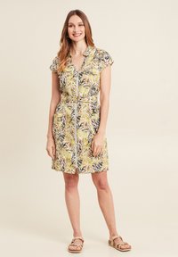 Kleid mit kurzen Ärmeln und Reißverschluss vorne, mit einem gelb-grünen Blattmuster auf leichtem Stoff und einem taillierten Gürtel.
