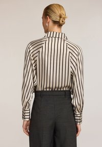 Blusa di seta crema con strisce verticali scure, con colletto e polsini abbottonati, abbinata a pantaloni scuri su misura. Texture liscia.