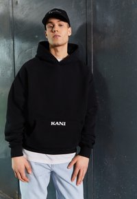 Black hooded sweatshirt with a front pocket, featuring white "KANI" text. Worn over a white shirt and paired with light blue jeans.