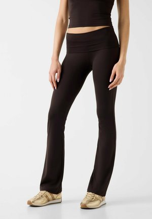 Vrouw draagt zwarte uitlopende legging, zwart crop top en beige sneakers, staand tegen een effen achtergrond.