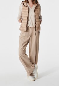 Beige puffer-väst med huva och svart dragkedja, kombinerad med en ljusbeige tröja och lösa, ljusbeige byxor, bärs med vita sneakers.