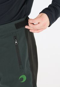 Schwarze und grüne Outdoor-Hosen mit strukturierter Oberfläche, ausgestattet mit einem Klettverschlussbund, einer Reißverschlusstasche und einem grünen Logo am Saum.