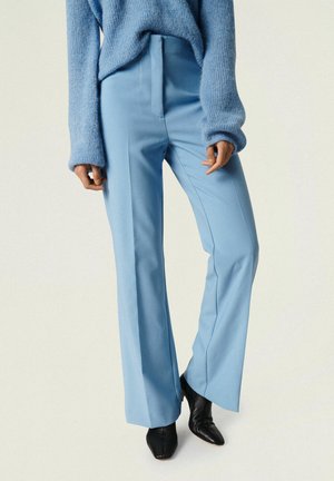 Kobieta ubrana w jasnoniebieskie, szyte na miarę spodnie o szerokich nogawkach, luźny, niebieski sweter z dzianiny i czarne botki z ostrym noskiem, stojąca na jednolitym tle.