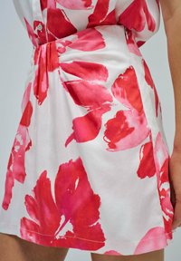 Primo piano di un vestito floreale con grandi e vivaci fiori rossi su uno sfondo chiaro, che mette in evidenza la texture del tessuto e una vita plissettata.