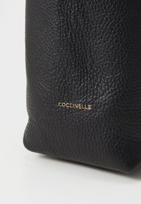 Black leather bag with a textured finish. Features embossed brand name "Coccinelle" in gold. Compact, rectangular shape.