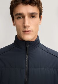 Granatowa kurtka z wysokim kołnierzem, wbudowanym zamkiem błyskawicznym i pikowaną strukturą. Posiada kontrastowe szwy oraz subtelny połysk.