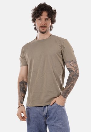 Uomo con capelli ricci che indossa una t-shirt marrone e jeans blu, che mostra tatuaggi dettagliati su entrambi gli avambracci.