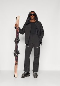 Zwarte skijas met een capuchon, zwarte basislaag, bijpassende broek met ritssluitingen, en skibotten. Houdt skies vast met bindingen.