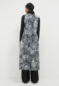 Donna che indossa un lungo gilet a spirale bianco e nero sopra un outfit nero, con un foulard nero e orecchini d'argento, vista di spalle.