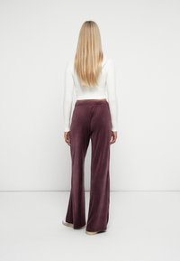 Frau mit langen blonden Haaren steht mit dem Rücken zur Wand, trägt ein weißes, langärmliges Crop-Top und lila weit geschnittene Hose sowie beige Schuhe.
