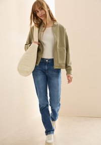 Chaqueta verde oliva con cremallera y dos bolsillos frontales, combinada con jeans azules y zapatillas blancas, llevando un bolso de hombro acolchado en color crema.