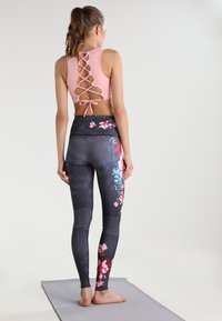 Vaaleanpunainen urheilullinen crop-top, jossa on ristikkäiset hihnat takana ja solmukoriste, yhdistettynä istuviin mustiin legginsseihin, joissa on kukkakuviota ja tekstuurisia yksityiskohtia.