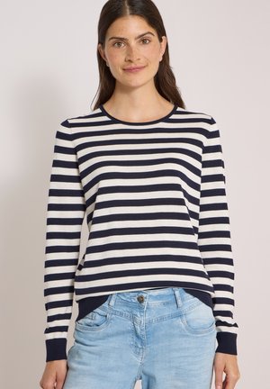 Jeune femme aux longs cheveux bruns portant un t-shirt rayé marine et blanc à manches longues et un jean bleu clair sur un fond uni.