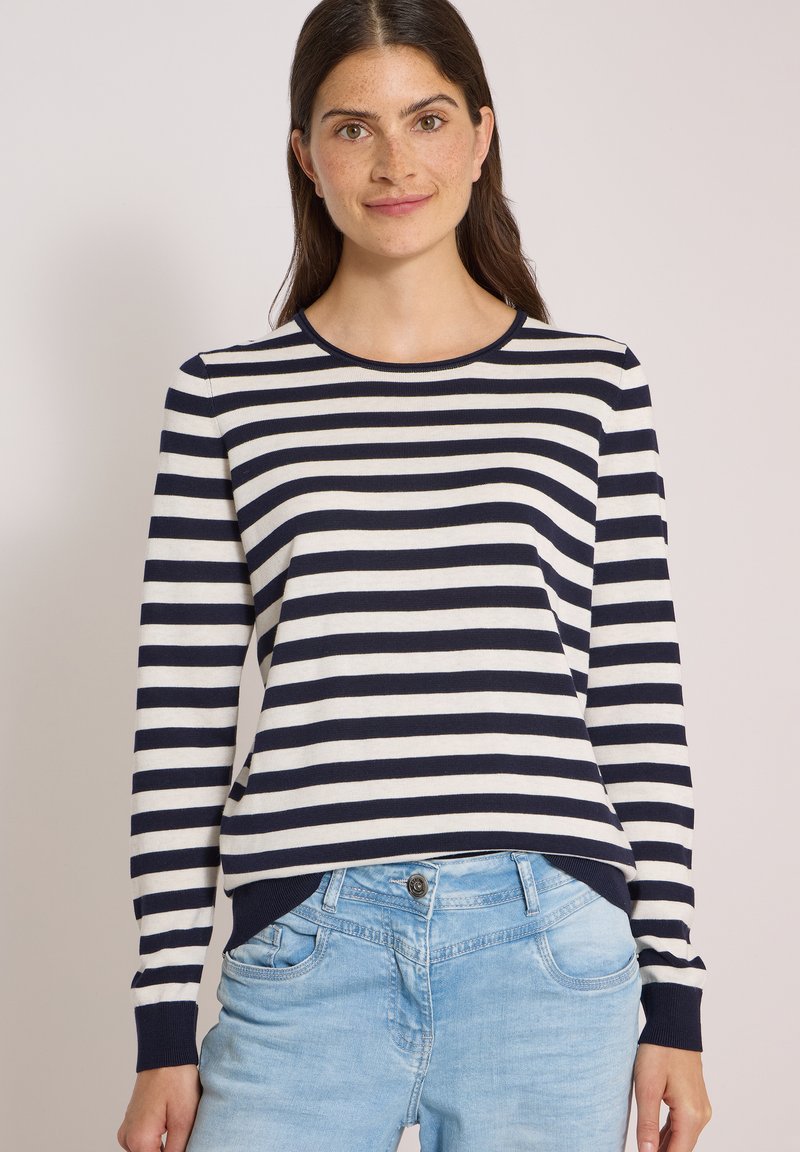 Jeune femme aux longs cheveux bruns portant un t-shirt rayé marine et blanc à manches longues et un jean bleu clair sur un fond uni.