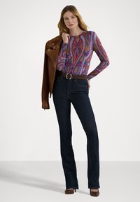 Hosszú ujjú, mintás felső lila, kék és narancssárga színekben, sötétkék, bővülő farmerral, barna bőrkabáttal és barna magassarkú cipővel párosítva.