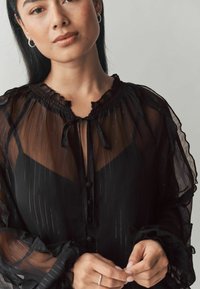 Blusa nera trasparente con strisce verticali, scollatura e polsini arricciati, bottoni frontali e un dettaglio con fiocco al collo. Tessuto morbido e leggero.