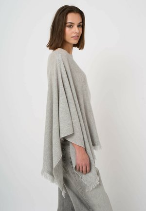 Kobieta o brązowych włosach do ramion, ubrana w jasnoszary ponczo z frędzlami na jasnoniebieskich dżinsach, stojąca na tle jednolitego białego tła.