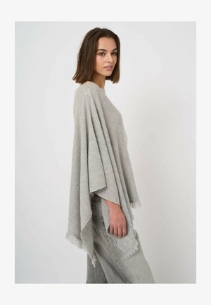 Vrouw met halflang bruin haar die een lichtgrijze poncho met franjes draagt over lichtgewassen spijkerbroek, staand tegen een effen witte achtergrond.